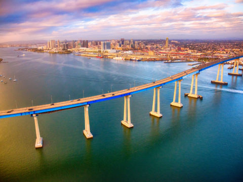 Aerial view of Coronado Bridge with vehicles, boats, and the San Diego skyline in the distance