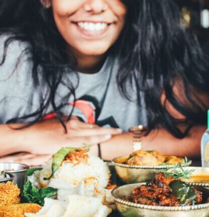 A smiling woman sits at a table filled with Indian dishes and a milkshake.