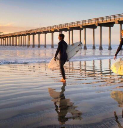 Two surfers carrying boards along the wet sand at sunrise near Scripps Pier.