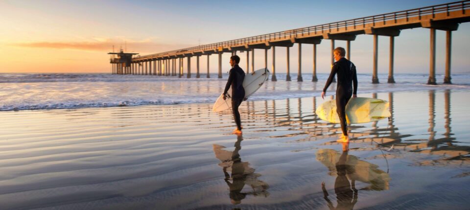 Two surfers carrying boards along the wet sand at sunrise near Scripps Pier.