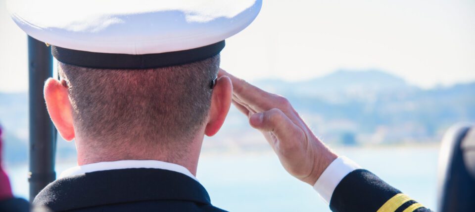 A uniformed sailor gives a salute while facing the ocean in bright daylight.