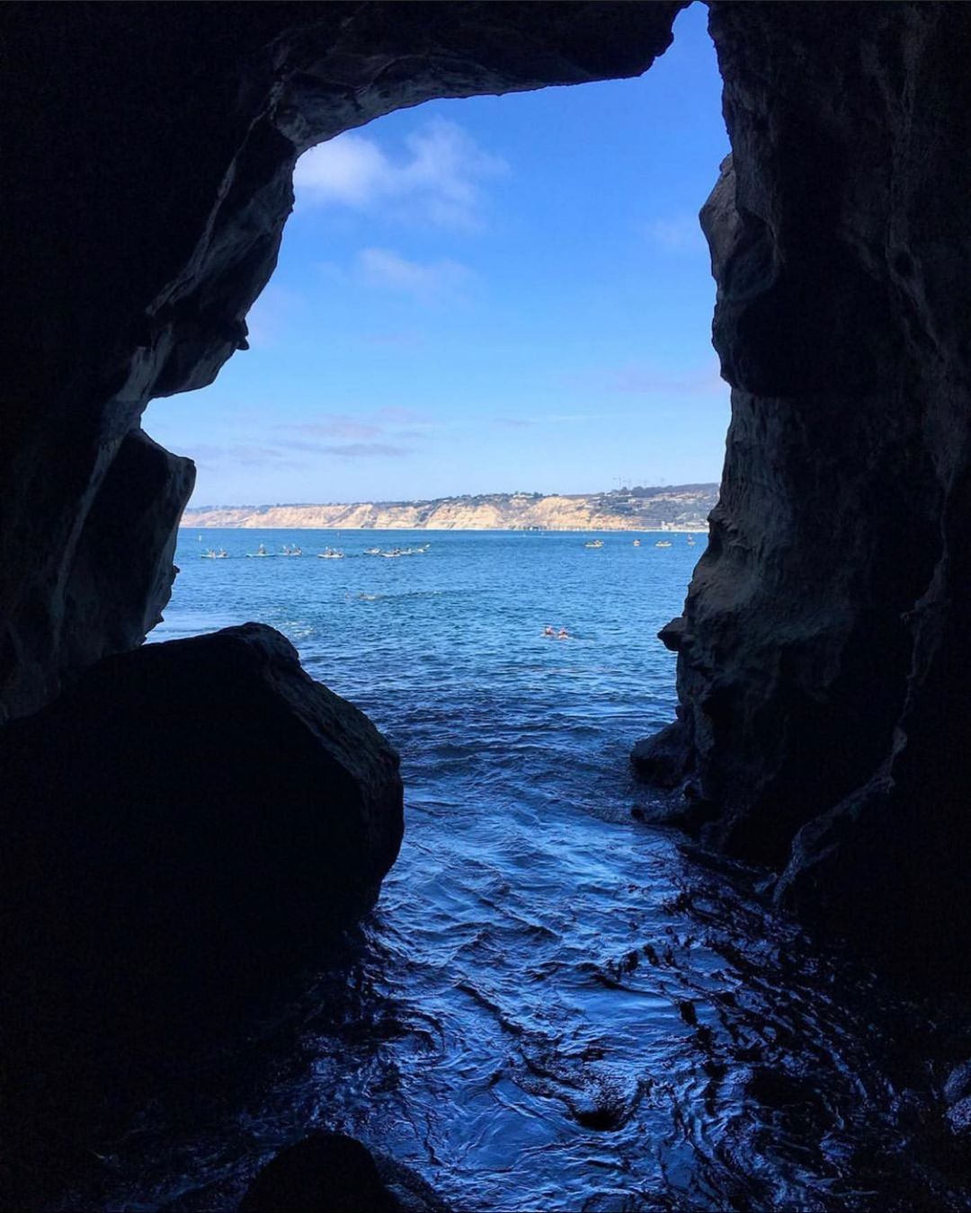 Ocean view from inside a dark sea cave looking out toward kayakers and coastal cliffs.