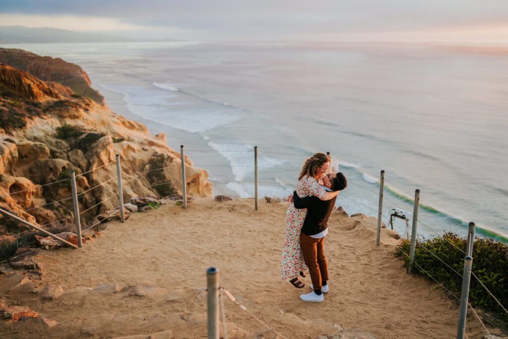 A couple embracing at the edge of a sandy cliff trail overlooking the ocean at Torrey Pines.