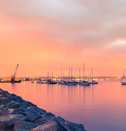 Sailboats anchored on calm water at sunset with the San Diego skyline glowing in the distance.