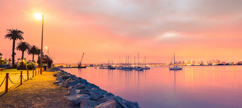 Sailboats anchored on calm water at sunset with the San Diego skyline glowing in the distance.