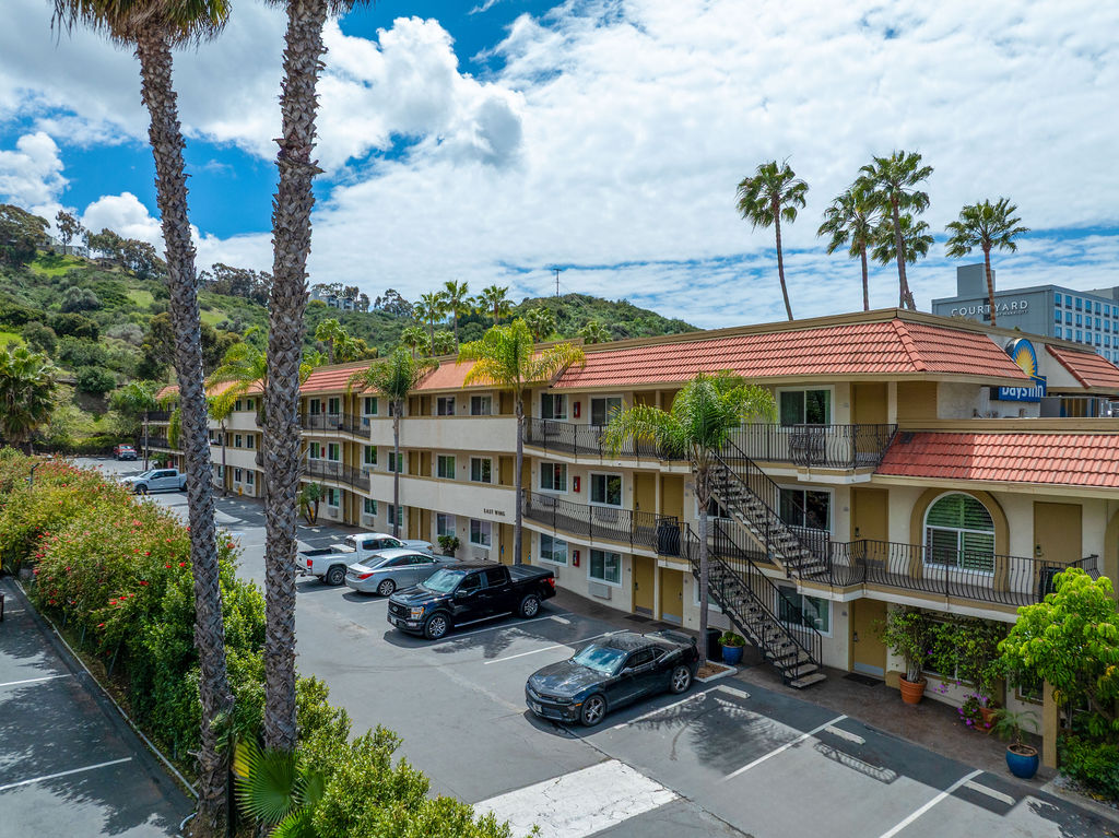 Hotel with red-tiled roof, balconies, palm trees, parking lot.